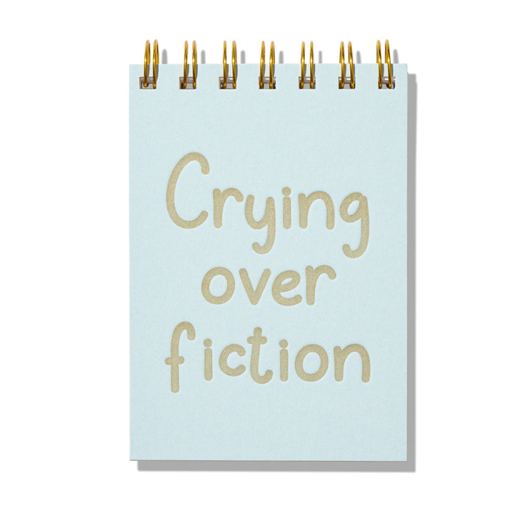 Blue mini jotter with letterpress text “Crying Over Fiction” and gold spiral binding.