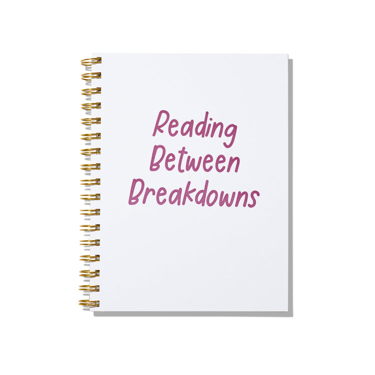 Large letterpress notebook with magenta text “Reading Between Breakdowns” on white cover with gold spiral binding.