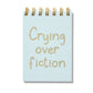 Blue mini jotter with letterpress text “Crying Over Fiction” and gold spiral binding.
