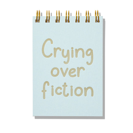 Blue mini jotter with letterpress text “Crying Over Fiction” and gold spiral binding.
