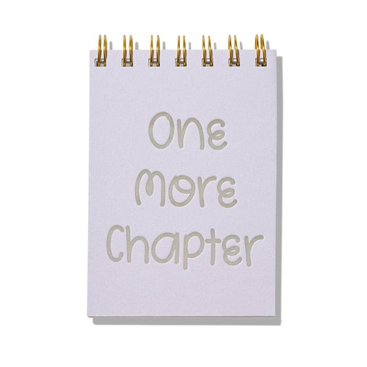 Purple mini jotter with letterpress text “One More Chapter” and gold spiral binding.