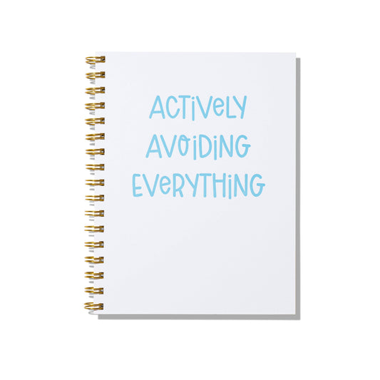 Large letterpress notebook with blue text “Actively Avoiding Everything” on white cover with gold spiral binding.
