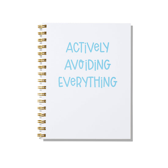 Large letterpress notebook with blue text “Actively Avoiding Everything” on white cover with gold spiral binding.
