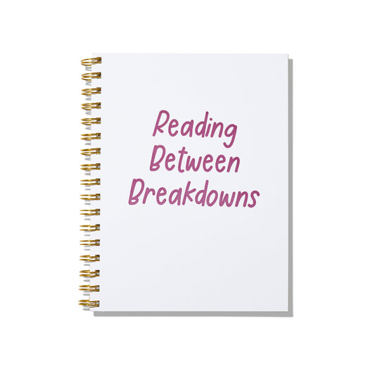 Large letterpress notebook with magenta text “Reading Between Breakdowns” on white cover with gold spiral binding.