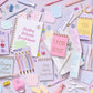 Assorted stationery items including notebooks, pens, and magnets on a light purple background